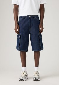 Pantaloni scurți cargo din denim de culoare albastru închis, cu buzunare laterale și o croială lejeră, asortați cu un tricou alb și teniși gri.