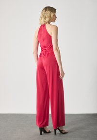 Rød jumpsuit med halterneck, vide ben og elastisk midje. Glatt tekstur og minimalistisk ryggdetalj. Svarte hælhæler.