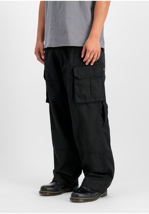 Person, die schwarze Cargohosen mit großen Seitentaschen, schwarze Lederschuhe mit gelber Naht und ein graues Hemd trägt, stehend vor weißem Hintergrund.