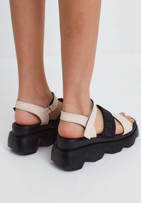 Beige szandál fekete rugalmas szalaggal, amelyen a "FASHION" felirat található, vastag fekete platform talppal, állítható bokapántokkal és sima textúrával.