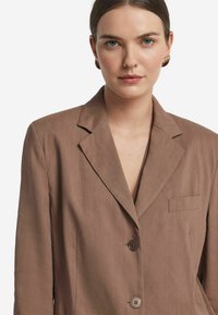 Brązowa marynarka wykonana z gładkiego materiału, z klapami w kształcie litery notched, pojedynczą kieszenią na piersi i dwoma ciemnymi guzikami z przodu.