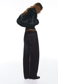 Veste courte en fausse fourrure noire avec manches larges, associée à un jean en denim noir ample et taille haute. Le mannequin est de profil, dévoilant le ventre.