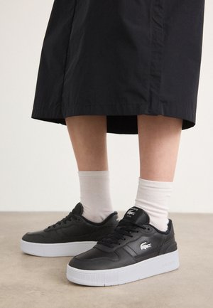 Personne portant des baskets Lacoste noires avec des semelles blanches, des chaussettes blanches et une jupe noire, debout sur un sol clair.