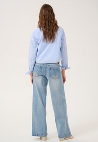 Camicia azzurra chiara con bottoni, maniche lunghe, abbinata a jeans a gamba larga e scoloriti. La vista posteriore mostra una vestibilità rilassata e dettagli minimi.