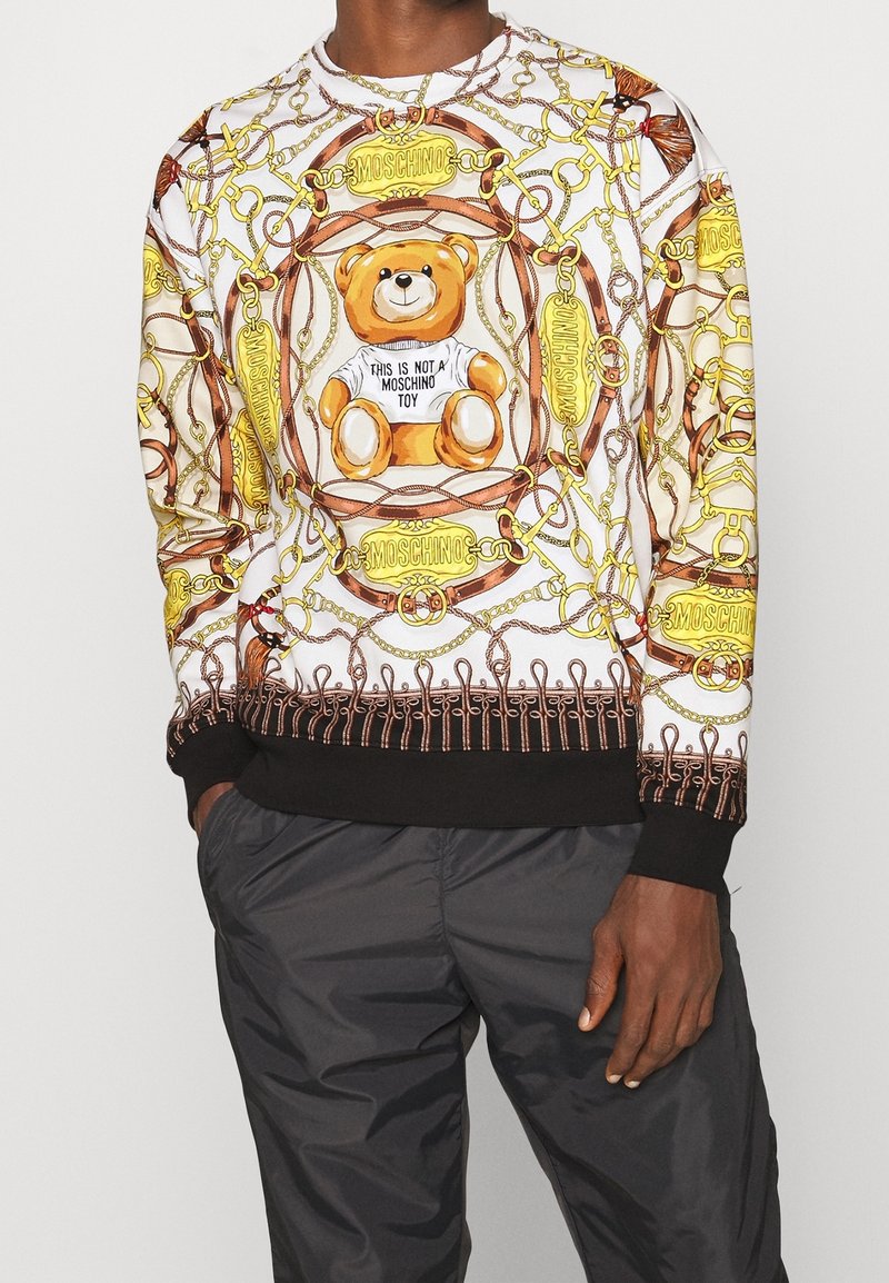 Sweat-shirt avec un motif d'ours en peluche portant une chemise où est inscrit « This is not a Moschino toy », entouré de chaînes en or et de logos sur tissu blanc.