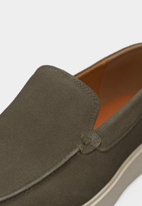 Πράσινο ελιάς σουέτ παπούτσι slip-on με ταμπά εσωτερική επένδυση και λευκή καουτσούκ σόλα, κοντινή θέα του επάνω μέρους και των ραφών στο πλάι.