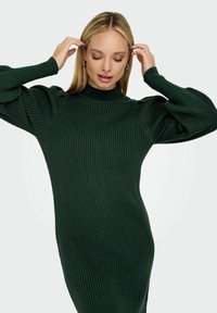 Robe en tricot côtelé vert foncé avec un col haut, des manches ballon et une silhouette ajustée. Présente un tissu texturé dans tout l'ensemble.