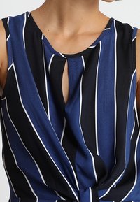 Marinblus och svart ärmlös topp med vita vertikala ränder, med ett nyckelhålsurringning och en knutet detalj vid midjan. Slät tygtextur.