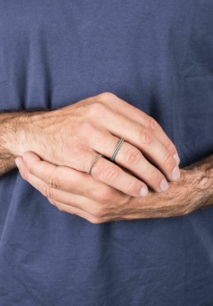 Dos anillos en una mano, uno de plata con una banda texturizada y el otro una banda delgada y lisa. La mano tiene piel de color marrón claro y una cantidad moderada de vello.