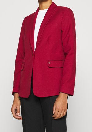 Person iført en tætsiddende rød blazer med klaplommer over en hvid top og sorte bukser, stående mod en ensfarvet lys baggrund.