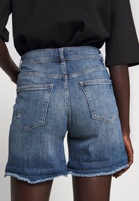 Person som bär mellanblå jeansshorts med fransiga kanter och en svart lös skjorta, sedd bakifrån mot en enkel bakgrund.