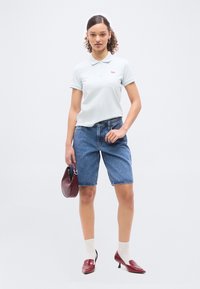 Γυναίκα που φοράει λευκό πόλο μπλουζάκι, σορτς από τζιν μέχρι το γόνατο, λευκές κάλτσες, μπορντό ψηλοτάκουνα και κρατά μια μικρή μπορντό τσάντα χειρός.