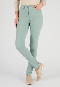 Jeans ajustados de color verde claro con una textura suave, cintura alta, bolsillos delanteros y cierre de botón único, combinados con zapatillas beige.
