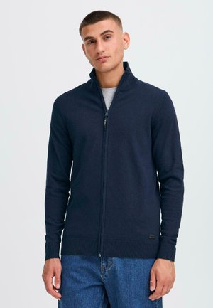 Mornarska zip-up jopica iz mehkega materiala, z ribanim robom in manšetami, visokim ovratnikom ter nevsiljivim logotipom na spodnjem levem delu. Oblačena čez sivo majico.