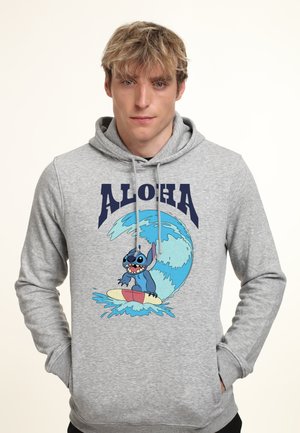 Junger Mann trägt einen grauen Kapuzenpullover mit dem Aufdruck "ALOHA" und einer blauen Cartoon-Figur, die auf dem vorderen Design eine große Welle surft.