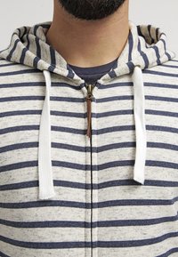 Felpa con zip a righe in crema e blu navy, dotata di un cordino beige, zip a contrasto e un tessuto morbido e textured.
