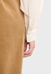 Tessuto in corduroy tan con cordoni verticali e una blusa di colore crema con maniche plissettate e polsino con bottoni, mano appoggiata di lato.