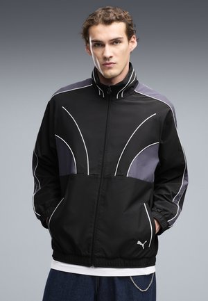 Junger Mann mit heller Haut und lockigem Haar, der eine schwarze und graue Puma-Zip-Up-Jacke mit weißen Paspeln trägt, steht vor einem grauen Hintergrund.