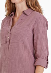 Woman wearing a mauve textured long-sleeve shirt with rolled sleeves, two chest pockets, and a partially buttoned collar.