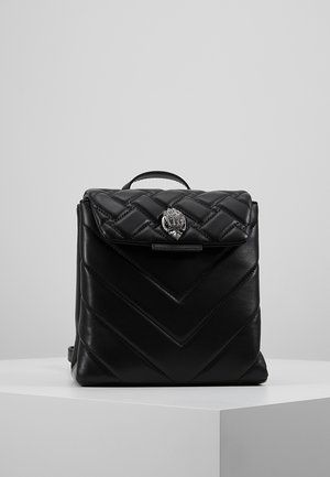 Sac à dos en cuir matelassé noir avec rabat supérieur, fermoir argenté en forme de coquillage et une seule poignée sur un piédestal blanc.