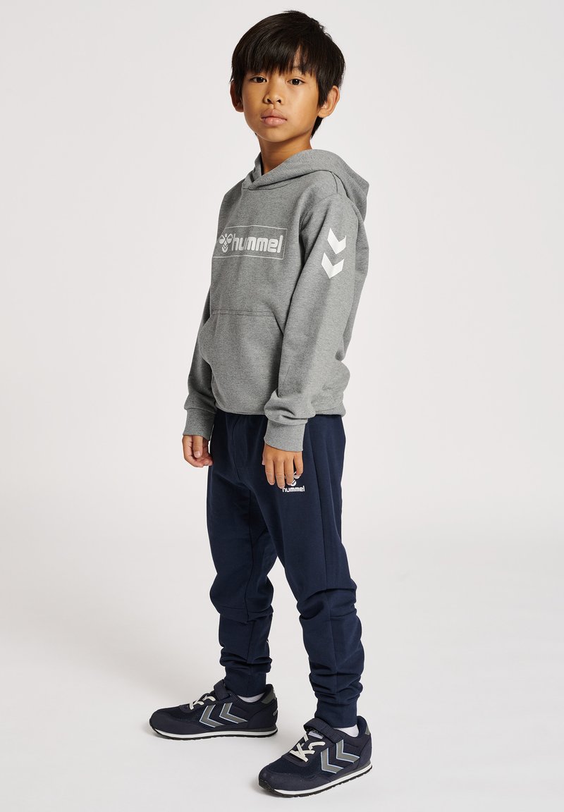 Garçon debout portant un sweat à capuche gris Hummel, un pantalon de jogging bleu marine Hummel et des baskets bleu marine, légèrement tourné vers la gauche sur un fond uni.