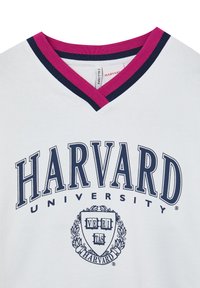 T-shirt bianco in cotone con scollo a V navy e rosa. Presenta la scritta "HARVARD UNIVERSITY" in grassetto di colore scuro e un emblema con uno scudo e una corona d'alloro.