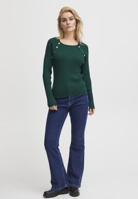 Maglione verde a costine con dettagli a bottoni sulle spalle, abbinato a jeans a zampa d'elefante blu e stivaletti neri alla caviglia. Sfondo bianco semplice.