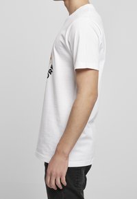 Mladý člověk oblečený v bílém tričku s krátkým rukávem a černých džínách, stojící v profilu na jednobarevném světlém pozadí.