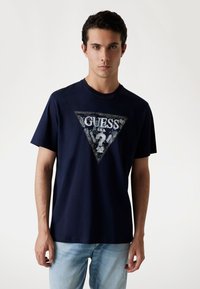 Dunkelblaues Baumwoll-T-Shirt mit einem dreieckigen, bedruckten Logo und komplizierten Designs sowie dem Wort "GUESS" in der Mitte. Kurze Ärmel.