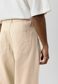 Beige spijkerbroek gezien van achteren, met een rechte pijp, twee achterzakken en een effen tailleband. De stof heeft een gladde textuur.