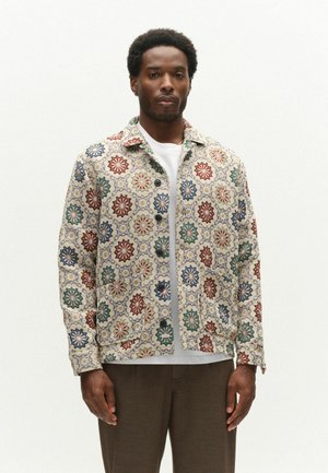 Mann trägt eine geknöpfte Jacke mit einem mehrfarbigen geometrischen Blumenmuster, darunter ein weißes Hemd und eine dunkelbraune Hose, steht vor weißem Hintergrund.