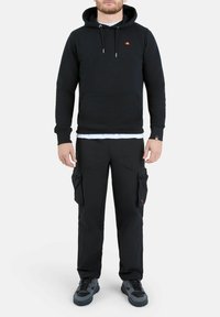 Uomo che indossa un hoodie nero con tasca frontale e pantaloni cargo neri con tasche laterali, in piedi contro uno sfondo chiaro e uniforme.