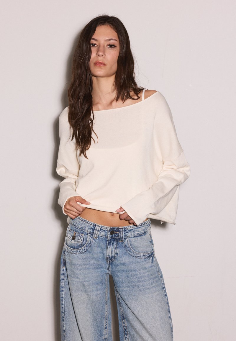 Ung kvinde med langt mørkt hår iført en løs off-white sweater og lyseblå bukser med vidde, står mod en ensartet lys baggrund.