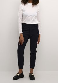 Vit långärmad topp med volangkrage, kombinerad med marinblå figursydda byxor med sidoglittrar. Svarta loafers med guldaccenter.