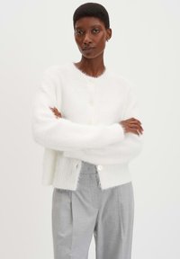 Weißer, flauschiger Cardigan mit Rundhalsausschnitt und zwei Knöpfen vorne, mit gerippten Bündchen und Saum. Kombiniert mit hellgrauen Hosen.