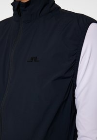 Marinblå ärmlös väst med hög krage, lätt material och en svart logotyp på bröstet, med rynkad textur längs ärmhålet.