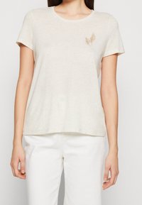 Camiseta de color beige claro hecha de una tela suave, con cuello redondo y mangas cortas, con un sutil diseño de hojas bordado en el pecho.