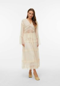 Robe longue beige clair avec accents en dentelle, tissu en tulle superposé, manches longues à volants et décolleté en V. Portée avec des chaussures beiges à talons blocs.