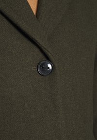 Manteau en laine vert olive foncé avec un seul bouton noir fermé près de la poitrine, porté par-dessus un vêtement intérieur marron.