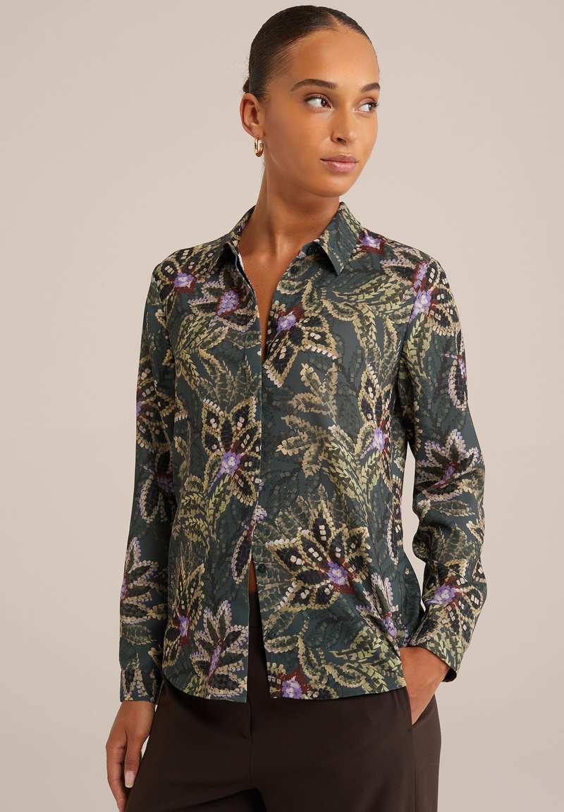 Knoopshirt met een donkergroene achtergrond met een bloemenpatroon in paars, geel en zwart. Lange mouwen en een klassiek kraag.