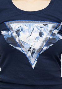 Μπλε πουκάμισο ναυτικού με μεγάλο τριγωνικό λογότυπο Guess που διαθέτει στρας και λευκό ανθισμένο μοτίβο στο κέντρο.