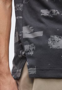 Μαύρο και γκρι υφασμάτινο μοτίβο σε πουκάμισο με πλαϊνή σχισμή, φορεμένο πάνω από μπεζ παντελόνι, που δείχνει το αριστερό χέρι και βραχίονα ενός ατόμου.