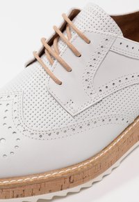 Chaussure en cuir blanc avec un design perforé, lacets marron clair et semelle en liège. Présente des coutures décoratives et un embout arrondi.