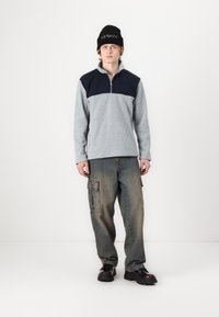 Pullover grigio in fleece con yoke blu navy, colletto zip e maniche lunghe, abbinato a pantaloni cargo in denim larghi e stivali neri.