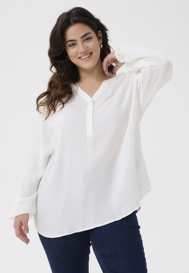 Weiße Bluse mit langen Ärmeln und strukturierten Oberfläche, V-Ausschnitt und Knöpfen vorne; kombiniert mit dunkelblauen Jeans.