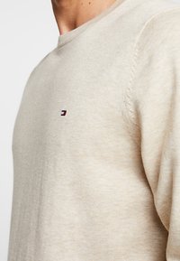 Beżowy dzianinowy sweter z okrągłym dekoltem, zdobiony małym haftowanym logo w kolorach granatowym, czerwonym i białym na piersi. Miękka tekstura.