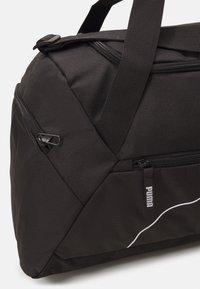 Crni sportski duffel torba s teksturiranim materijalom, istaknutim patentnim zatvaračima, dvostrukim ručkama i bijelim logotipom duž bočne strane.