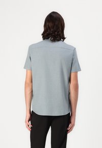 Camisa de manga corta en tejido gris claro con una textura sutil. Presenta un dobladillo recto y detalle en el yugo trasero. Combinada con pantalones negros.