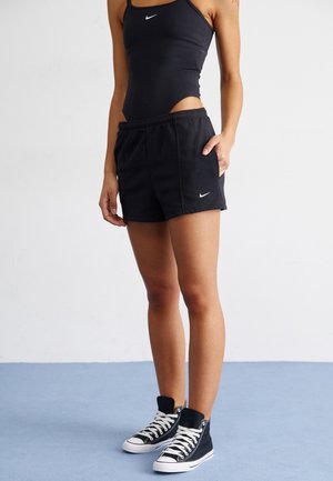 Femme portant un body noir sans manches Nike, un short Nike noir et des baskets montantes noires, debout sur un sol bleu.