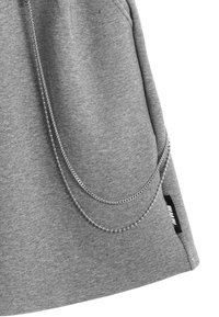 La tela del sudadera gris presenta un bolsillo con acentos de cadena metálica, detalle de costura lateral y una etiqueta de marca cerca del dobladillo.